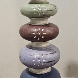 May include: A decorative, stacked wooden candle holder with a brown, yellow, green, burgundy, purple, and blue color scheme. Each section is a different color and has a white sunburst design. The base and top are brown.
