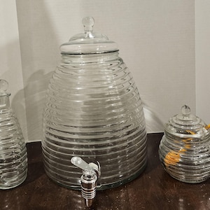May include: Clear glass beverage dispenser with a spigot, a matching jar, and a decorative bottle. The dispenser and jar have a ribbed design. The jar has a lid and a yellow spoon. The bottle has a stopper.