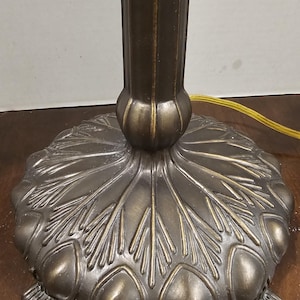 May include: Close-up of a bronze-colored lamp base with a detailed, floral-inspired design. The base features a round, textured surface with leaf-like patterns and small feet. The lamp's central column is also bronze, with a ribbed texture.