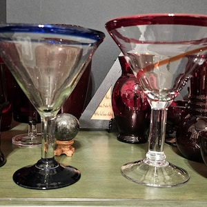 May include: Two clear glass margarita glasses with a blue and red rim. The glass on the left has a blue rim and the glass on the right has a red rim. The glasses are sitting on a wooden table.