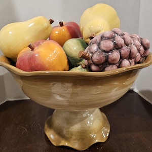 May include: A ceramic fruit bowl with a pedestal base, filled with colorful ceramic fruit. The bowl contains apples, pears, grapes, and other fruits in shades of yellow, red, green, and purple. The bowl has a light brown finish.