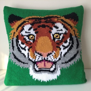 May include: A green knitted pillow with a tiger face design. The tiger has orange and brown stripes, black eyes, and a white mouth.