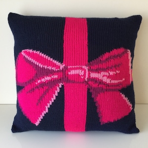 May include: A navy blue knitted cushion featuring a vibrant pink bow and vertical stripe design. The bow is centrally placed and has a detailed, textured appearance. The cushion is square-shaped.