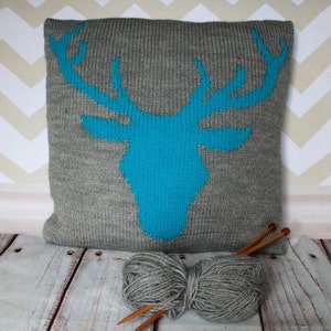 May include: A gray knitted pillow with a teal blue deer silhouette design. Two wooden knitting needles and a ball of gray yarn are in the foreground.