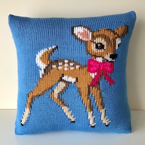 May include: A blue knitted pillow with a brown and white deer wearing a pink bow. The deer is facing the right side of the pillow.