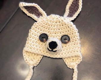 May include: A crocheted beige kangaroo hat with brown ears and black button eyes. The hat has a long, crocheted cord that ties under the chin.