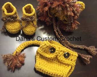 Op de afbeelding: Een gehaakt leeuwenkostuum voor een baby, bestaande uit een gele muts met bruin bont, een gele luierhoes met twee zwarte knopen en gele slofjes met bruine bies. Dana's Custom Crochet.