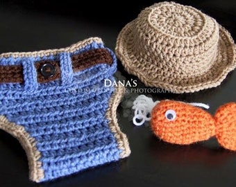 Puede incluir: Un conjunto de ropa de bebé de ganchillo con un pañal azul y marrón, un gorro de ganchillo de color beige y un pez de ganchillo naranja.
