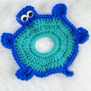 May include: A crocheted blue and teal turtle-shaped ring for newborn photography props. The ring has googly eyes and "Dana's Custom Crochet Photography" text.