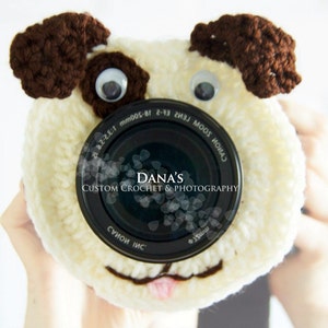 May include: A white crocheted camera lens cover shaped like a dog with brown ears and googly eyes. The lens cover has the text "Dana's Custom Crochet & Photography" printed on the lens.