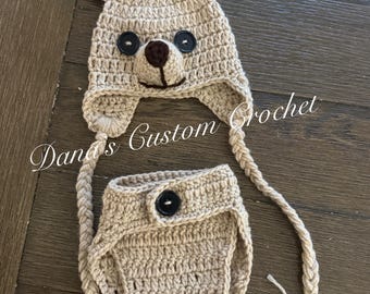 May include: A beige crochet hat and diaper cover set. The hat features two pointed ears with dark brown inner accents, two black button eyes, and a dark brown nose. Two long braided ties with tassels hang from the hat. The diaper cover has a black button.