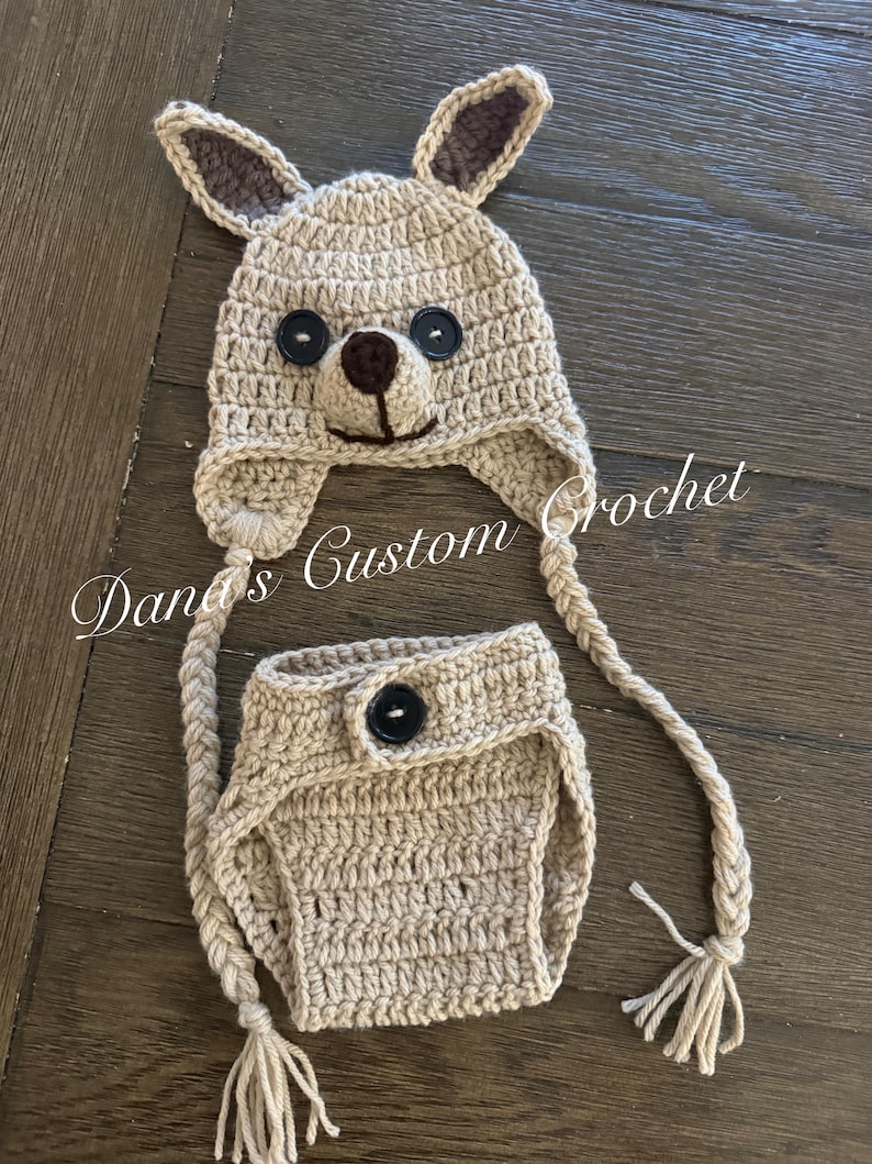 May include: A beige crochet hat and diaper cover set. The hat features two pointed ears with dark brown inner accents, two black button eyes, and a dark brown nose. Two long braided ties with tassels hang from the hat. The diaper cover has a black button.