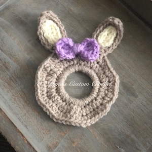 May include: A brown crocheted bunny ring with a purple bow. The ring is made of yarn and has a hole in the center.