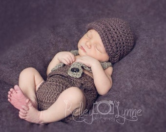 Puede incluir: Un bebé durmiendo con un gorro y un atuendo a juego de ganchillo marrón con un detalle de botón. El bebé está acostado sobre un fondo de tela morado.