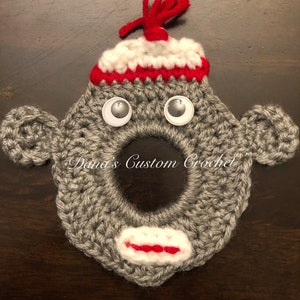 Op de afbeelding: Grijs gehaakt apen gezicht met een rode en witte muts, goggle ogen en een rode mond. De tekst "Dana's Custom Crochet" is zichtbaar in het midden van de afbeelding.