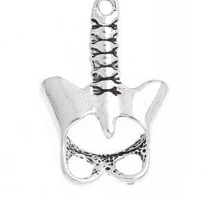 Puede incluir: Un dije plateado con forma de pelvis humana y columna vertebral inferior. El dije tiene un lazo en la parte superior para sujetarlo a un collar o pulsera. El dise&ntilde;o es detallado, con v&eacute;rtebras visibles y la estructura de la pelvis.