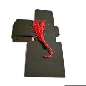 May include: Black favor box with a red tassel. The box is folded into a square shape with a flap on top. The tassel is attached to the flap and hangs down.