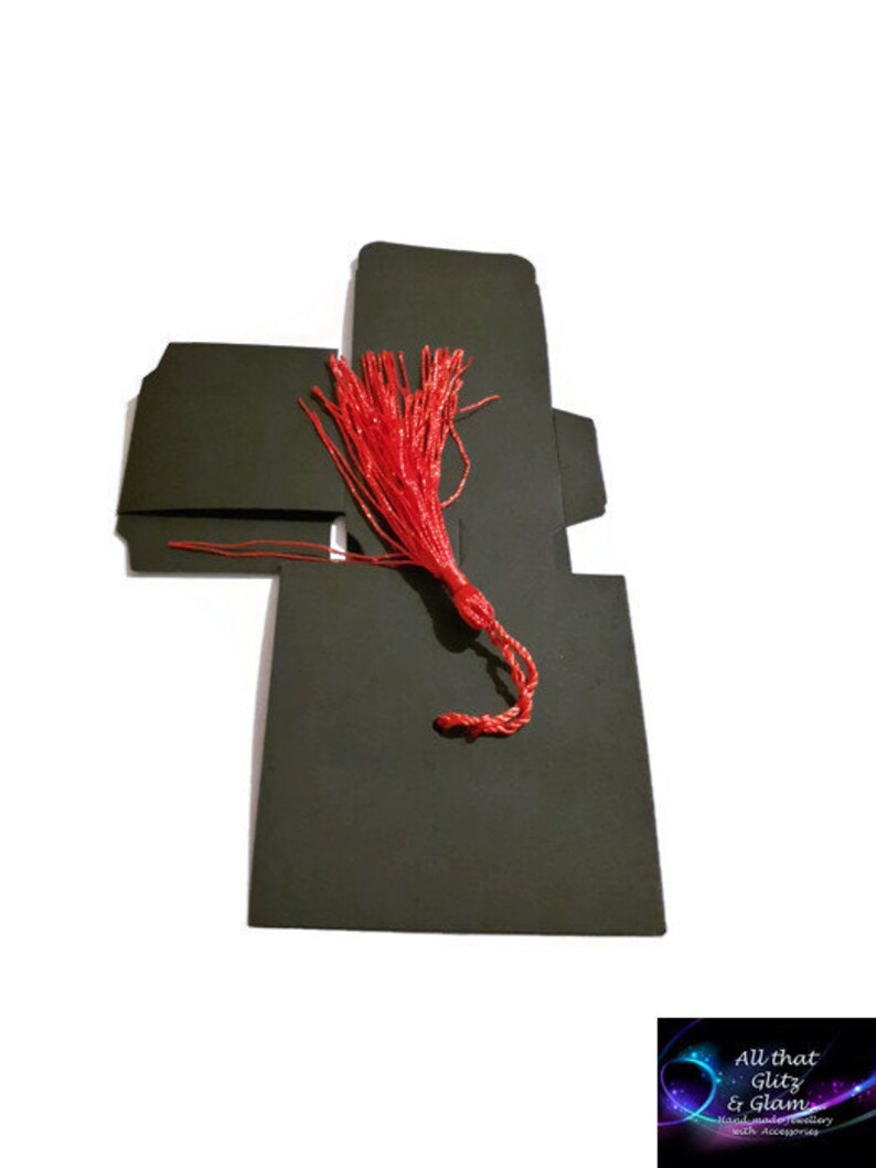 May include: Black favor box with a red tassel. The box is folded into a square shape with a flap on top. The tassel is attached to the flap and hangs down.