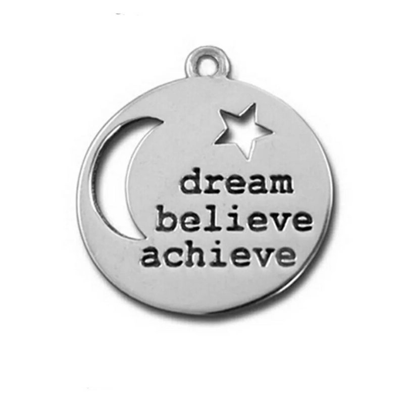 Puede incluir: Colgante plateado de forma circular. Presenta un recorte de media luna a la izquierda y un recorte de estrella en la parte superior derecha. Las palabras "dream believe achieve" est&aacute;n estampadas en el centro.