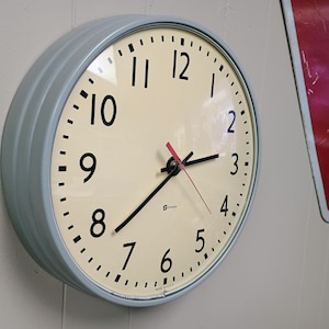 May include: A round wall clock with a light gray metal frame and a cream-colored face. The clock features black Arabic numerals, black hour and minute hands, and a thin red second hand. The word Simplex is printed in black on the clock face.
