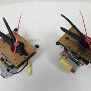 May include: Two clock mechanisms featuring brass-colored square plates, black hour and minute hands with a textured finish, and a thin red second hand. Each unit includes an attached electromagnetic coil component with yellow and copper wiring.