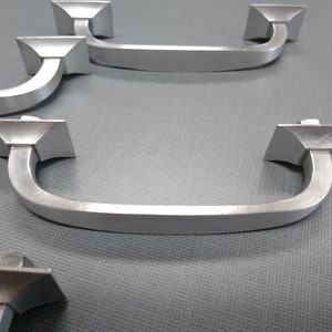 Puede incluir: Cuatro tiradores de armario plateados con un diseño cuadrado. Los tiradores tienen una forma curva y un acabado liso.