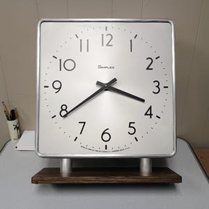 May include: A square Simplex wall clock with a white face, black numerals, and black hands. The clock is mounted on two silver cylindrical supports attached to a rectangular dark brown wooden base. The text Simplex is printed on the face.