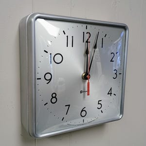 May include: A square wall clock with a silver frame and a white face featuring black Arabic numerals from 1 to 12. The clock has black hour and minute hands and a red second hand. The text Simplex and Made by Simplex Time Recorder Co. is visible.