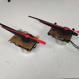 May include: Two clock mechanisms featuring brass-colored metal plates and cylindrical bases. Each unit has a long, textured black hour hand and a bright red minute hand attached to the center, with black wires extending from the rear of the components.