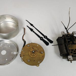 May include: Disassembled clock parts, including a silver metal casing, a circular brass mechanism with gears, black clock hands, and a black electrical component with wires. The parts are arranged on a white surface.