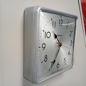 May include: A square, silver-colored Simplex clock with black numerals and hands, and a red second hand. The clock is mounted on a white wall. The time is approximately 10:35.