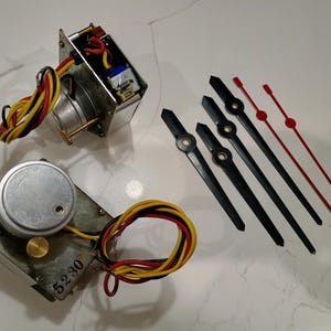 May include: Two silver-toned metal clock motor mechanisms with attached yellow, red, and black wiring bundles, accompanied by three black clock hands and two thin red clock hands, all featuring pointed tips and central mounting holes.