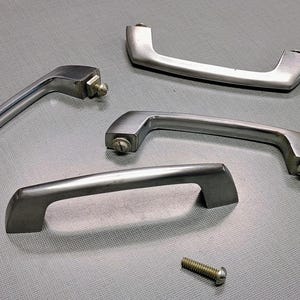 May include: Five silver-toned cabinet handles in various shapes, along with a single screw, are arranged on a gray surface. The handles have a brushed metal finish and are designed for easy grip. The hardware is suitable for kitchen or furniture projects.