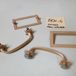 May include: Bronze cabinet hardware set, including two curved handles with hex nuts and two rectangular label holders. A handwritten tag reads "BRN-5 Bronze Shaw Walker."