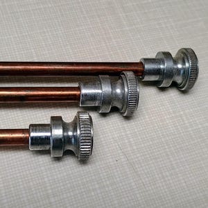 May include: Three copper-colored rods with silver-colored, decorative ends. The rods are of varying lengths and are arranged parallel to each other against a light gray background. The ends have a ridged design.