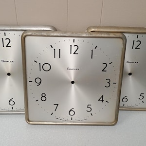 May include: Three silver square clocks with black numbers and hands. The clocks are all the same size and have a silver face. The clocks are all set to different times. The clocks are made by Simplex.