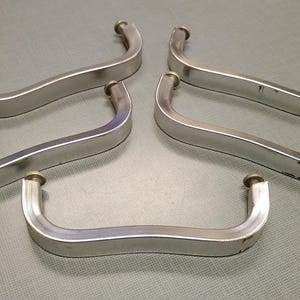 May include: Five silver-toned cabinet pulls with a curved, ergonomic design. Each pull has a rounded end on each side. The pulls are arranged on a gray surface, suggesting a hardware or home improvement theme.