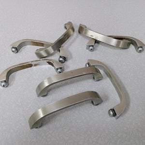 May include: A collection of six brushed metal cabinet handles. Each handle has a curved design with screw attachments on each end. The handles are silver and appear to be used.