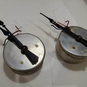 May include: Two circular silver-toned metal clock mechanisms featuring black metal hands. Each unit has exposed copper wiring and small screws on the top surface. The components appear to be vintage clock parts with a metallic finish and visible signs of age.