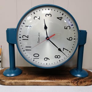 May include: A vintage Simplex clock with a teal frame and base, featuring a silver face with black numerals and hands. The clock sits on a wooden base. The second hand is red. A pencil holder with pencils is visible in the background.