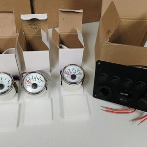 May include: A collection of marine gauges and a black control panel. The gauges have white faces with black markings and chrome bezels. The control panel has multiple switches and a voltage meter. Cardboard boxes are in the background.