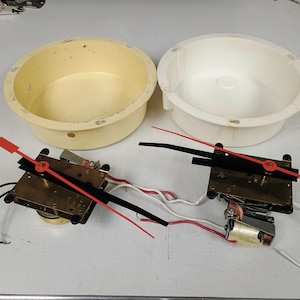 May include: Two round plastic clock housings, one cream and one white, paired with two clock mechanisms. Each mechanism features a brass-colored square base, a small motor, and two long, thin clock hands in contrasting red and black colors.