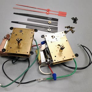 May include: Two brass-colored clock motor mechanisms with attached copper coils and electrical wiring. Included are two pairs of black clock hands, one pair of red clock hands, and a small collection of metal screws and fasteners for assembly.