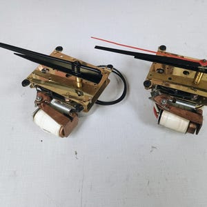 May include: Two clock movements featuring brass-colored metal frames, white cylindrical coils, and black hands. One movement includes a thin red second hand. These mechanical components are exposed, showing gears, wires, and mounting hardware for timepieces.