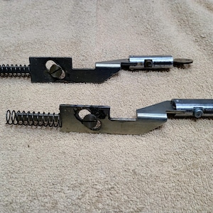 May include: Two metal latches with springs and keys. Each latch has a black, rectangular base with a circular hole and a silver-colored, angled arm. A coiled spring is attached to the base.