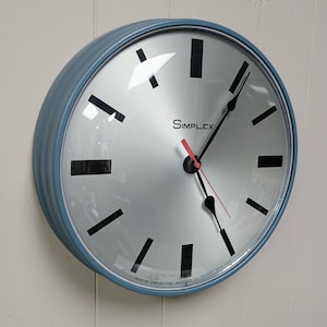 May include: A round wall clock with a dusty blue metal frame and a silver metallic face. The clock features black rectangular hour markers, black hour and minute hands, and a thin red second hand. The word SIMPLEX is printed in black.