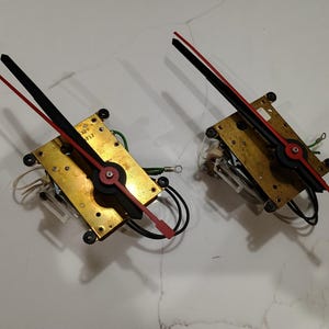 May include: Two clock mechanisms featuring rectangular brass-colored plates, black and red hands, and attached electrical wiring. Each unit has a central pivot point for the hands, exposed gears, and small mounting points at the corners of the plates.