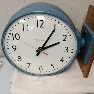 May include: A round, blue-cased Simplex wall clock with a cream-colored face, black numerals, and black hands. A thin red second hand is visible. The clock is mounted on a vertical wooden plank attached to a blue metal bracket on the right side.