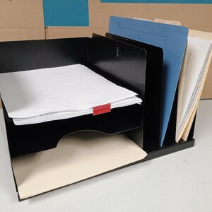 May include: Black metal desk organizer with multiple compartments. The organizer holds stacks of white paper, a red paper clip, and file folders in various colors. A practical office supply.