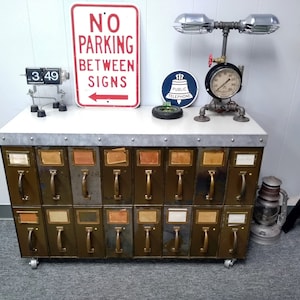 May include: A vintage metal cabinet with 18 drawers, each with a brass handle and a label. The cabinet is on wheels and has a gray metal top. There is a sign that says "No Parking Between Signs" and a clock that reads "3.49" on the top of the cabinet.